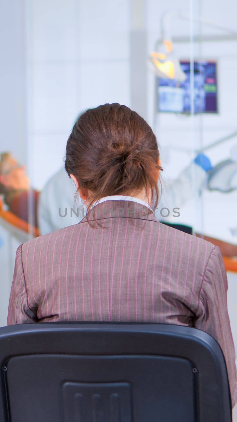 Back view of woman filling in dental form sitting on chiar in waiting room preparing for dental implants while doctor exemination patient in background. Crowded professional orthodontist office.