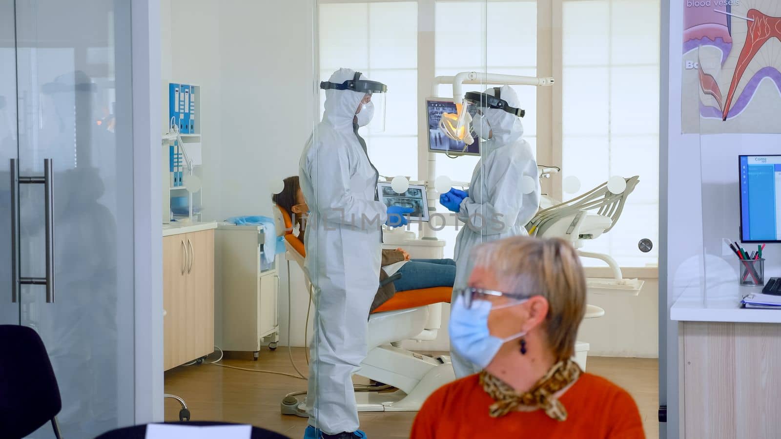 Doctors wearing full virus protection uniform standing in surgery room planning dental treatment while elderly patients waiting in reception keeping distance. Concept of new normal dentist visit