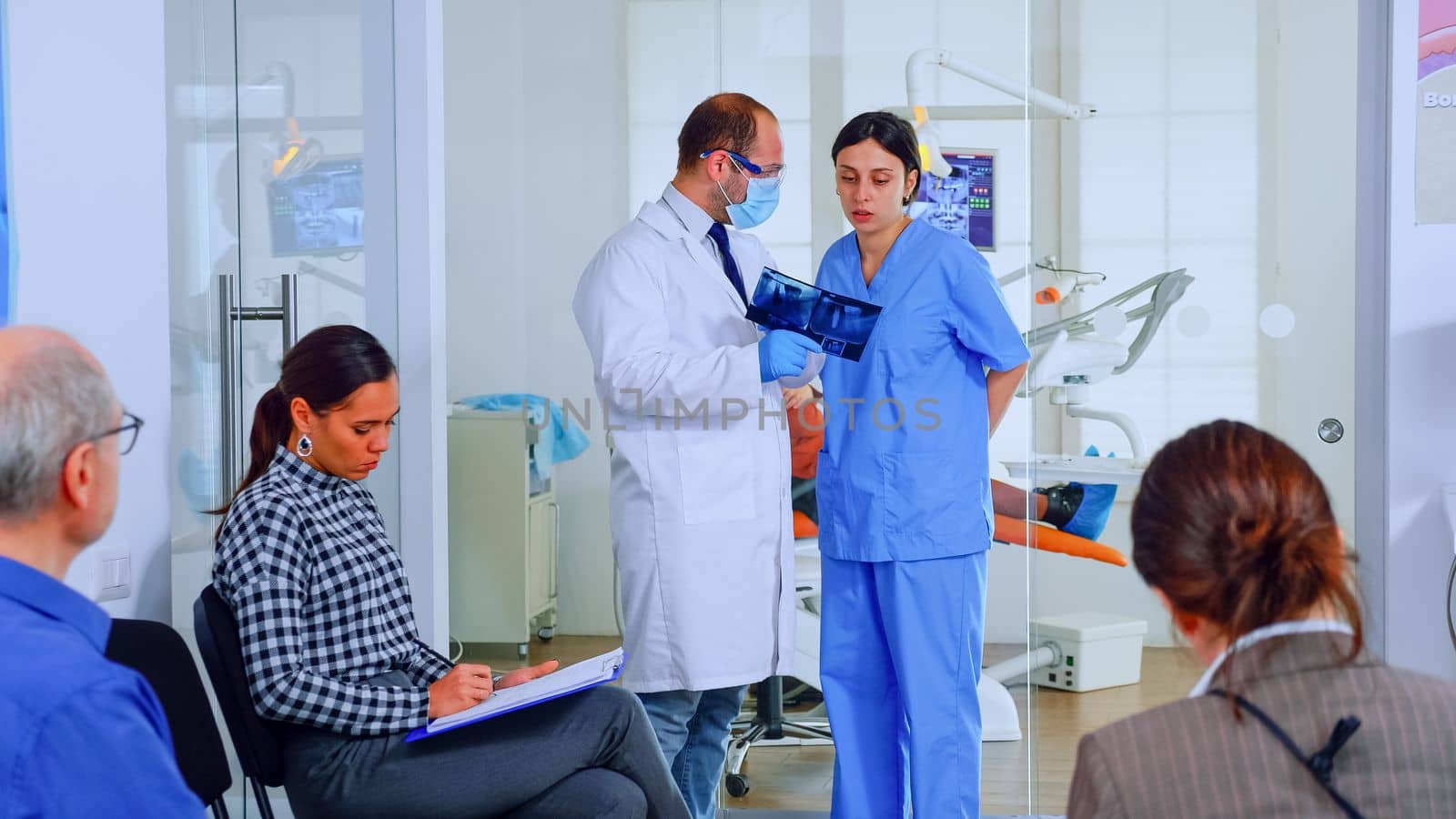 Dentist showing teeth x-ray reviewing it with nusre. Doctor and assistant working in modern crowded stomatological clinic, patients sitting on chairs in reception filling in dental forms and waiting