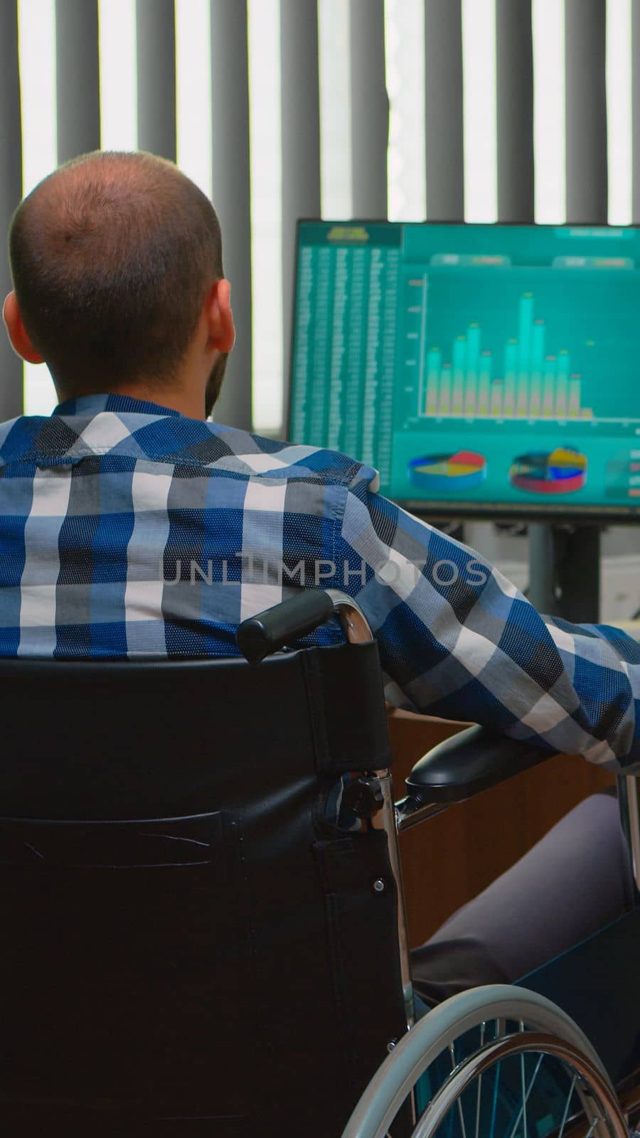 Disabled entrepreneur sitting in wheelchair immobilized analysing financial economy statistics on computer in business office discussing with colleague. Handicapped businessman using modern technology