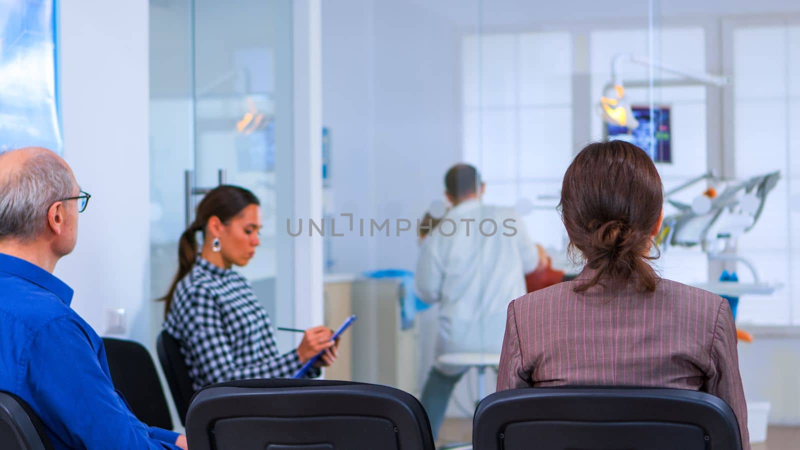 Back view of woman filling in dental form sitting on chiar in waiting room preparing for dental implants while doctor exemination patient in background. Crowded professional orthodontist office.