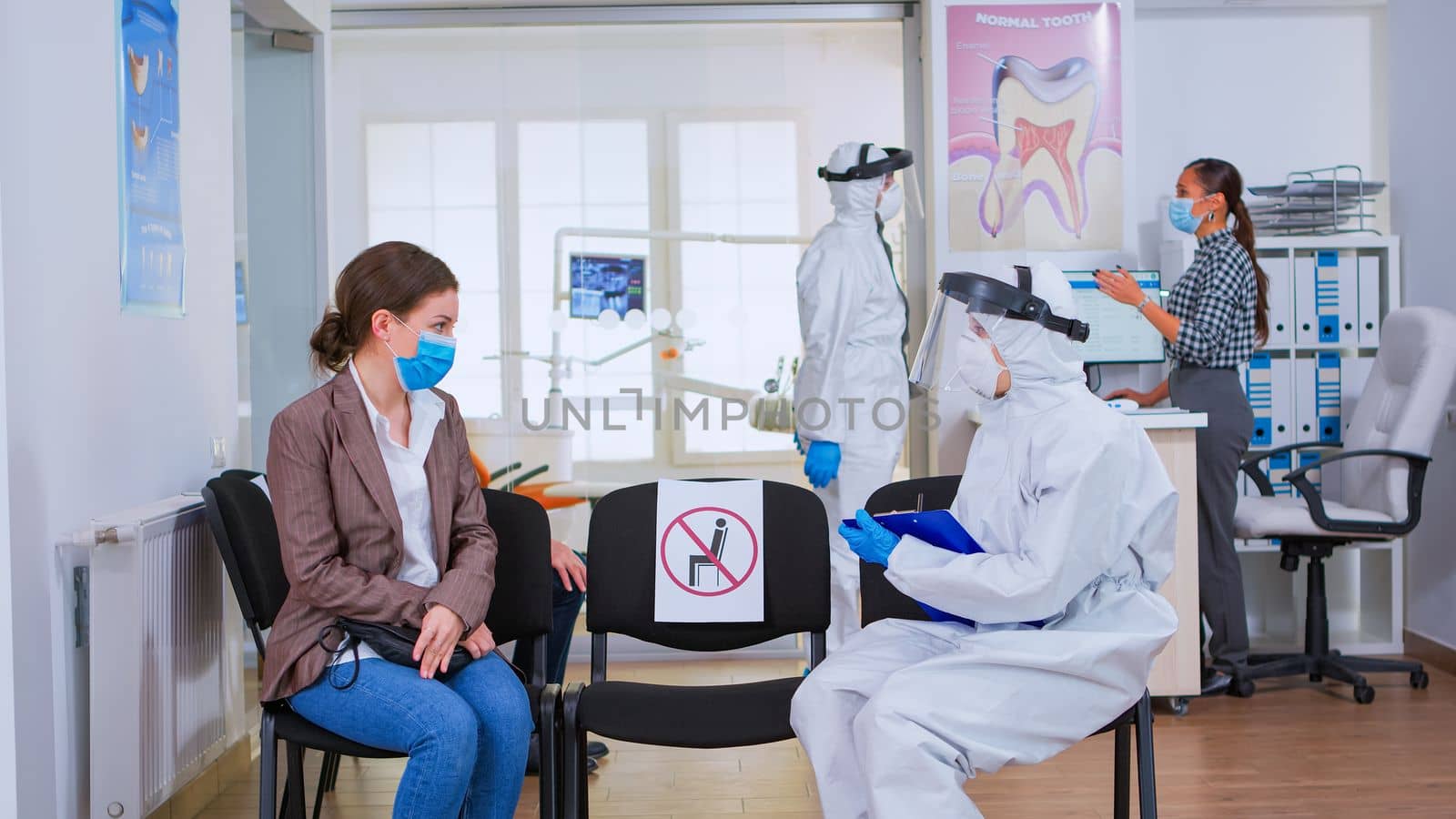 Dentist nurse with protection equipment talking with patient before consultation during covid-19 epidemic sitting on chairs in waiting area keeping distance. Concept of new normal dentist visit.