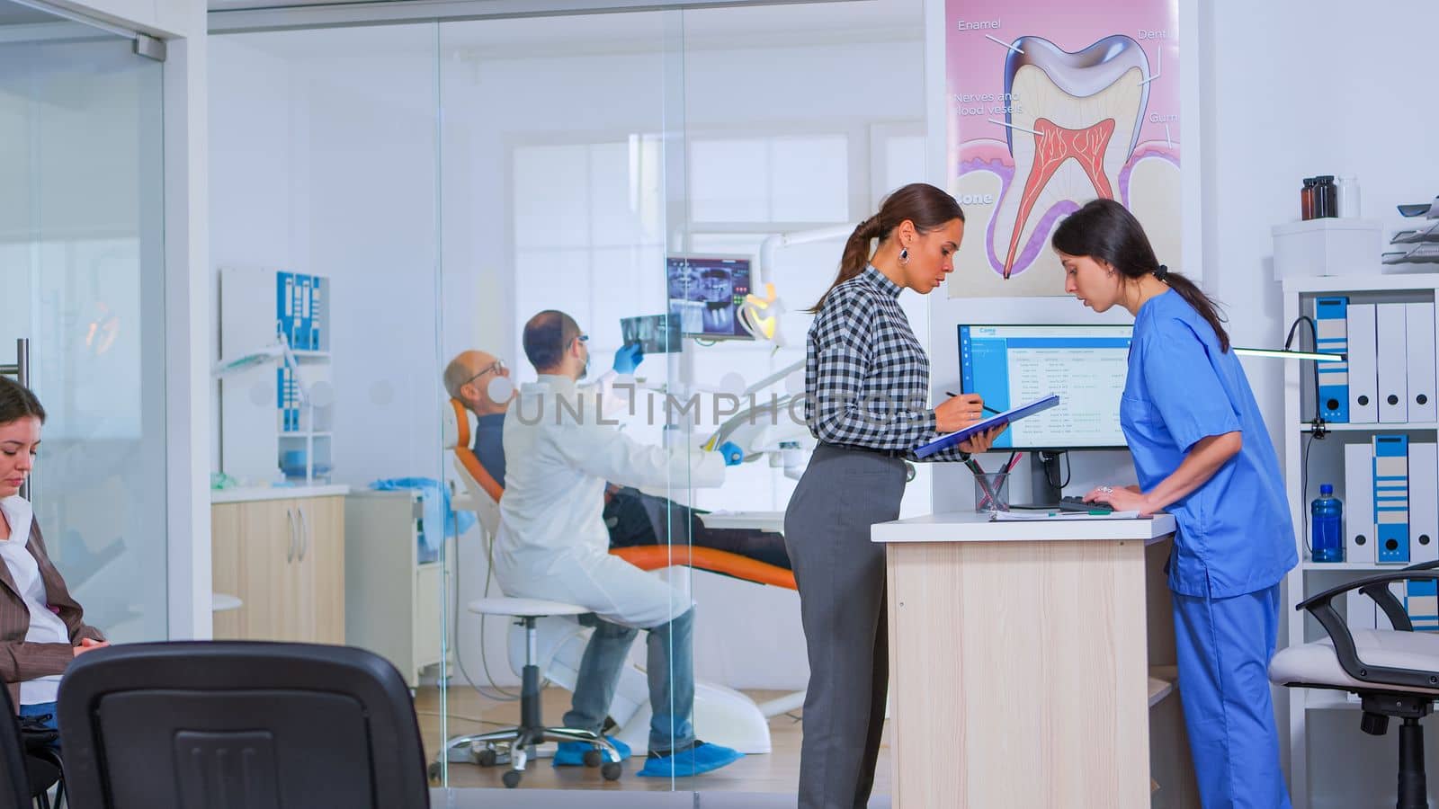 Professional dentist asking nurse for dental x-ray before examining patient while people waiting in reception area of modern stomatological clinic. Dental nurse typing on computer making appointments.