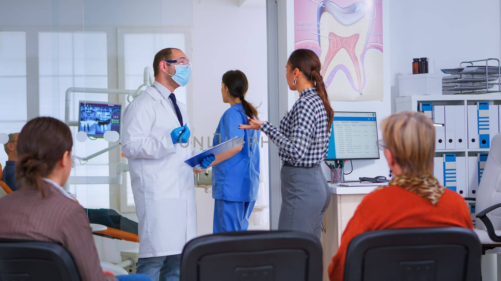Doctor taking notes on clipboard about woman dental problems standing in waiting area, while nurse preparing tools for next patient in consultation room. Dentist explaining medical procedure.