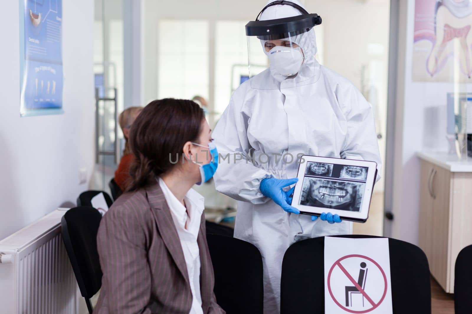 Stomatologist in protective suit pointing on digital x-ray of tooth explaining to patient treatment using tablet in covid-19 pandemic. Medical team wearing face shield, coverall, mask and gloves.