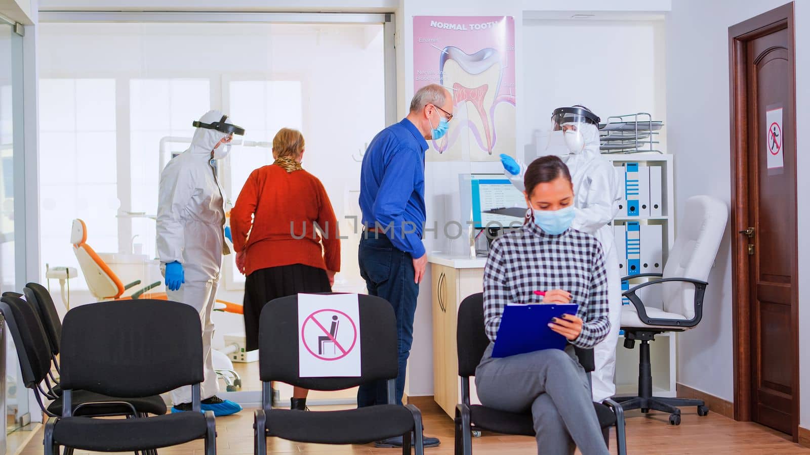 Dental assistant with coverall against coronavirus giving form to fill out after measuring patient temperature. Patient with face protection mask writing on registration document in stomatomoly clinic