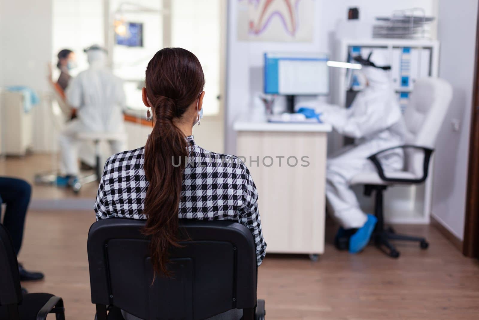 Woman patient in dentistiry office waiting for diagnosis in dental clinic corridor. Dentist and staff dressed in ppe suit consulting patient. Filling health form.