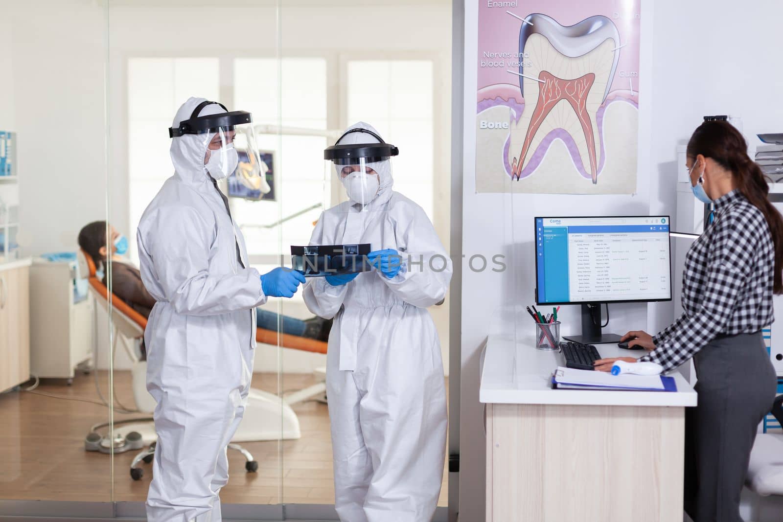 Stomatology team dressed up in ppe suit during global pandemic with coronavirus in dental reception holding patient x-ray, keeping social distancing. Receptionist with face mask as prevention.