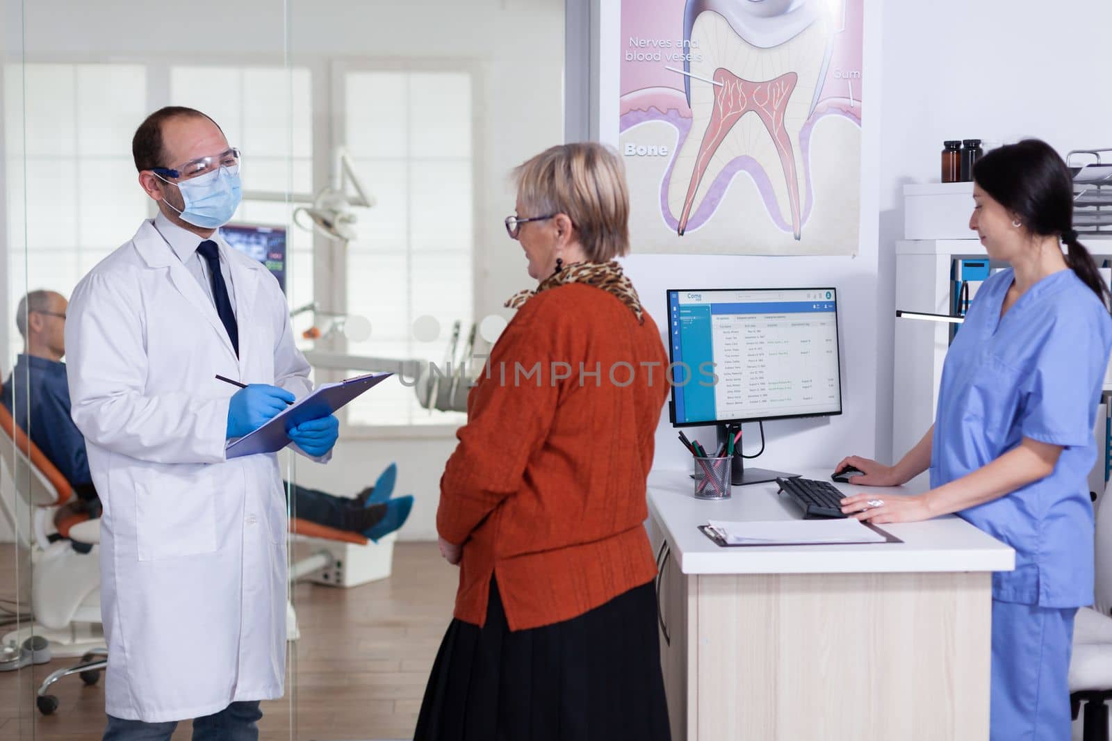 Dentist doctor with face mask explaining diagnosis to senior woman patient in stomatology waiting area hallway. Elderly man sitting on chair for teeth treatment.