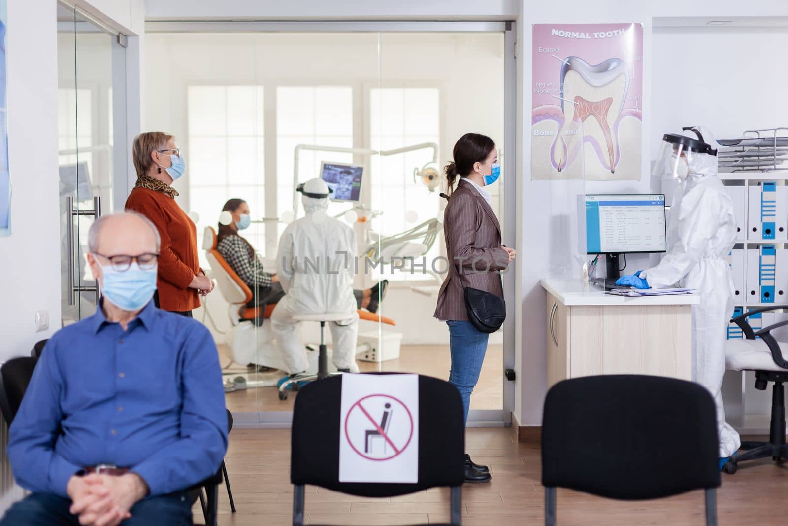 Patients with protection mask waiting in stomatology reception with new normal, staff wearing ppe suit. Woman checking appoiment with nurse coming for dental control during coronavirus pandemic