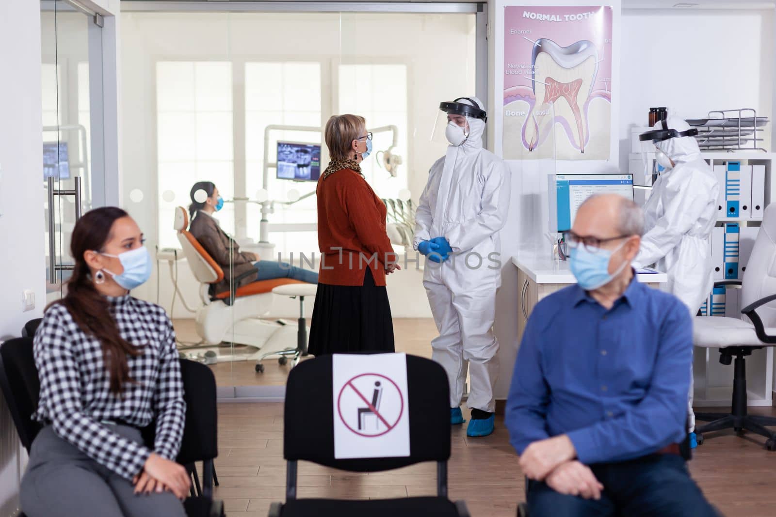 Senior woman patient in stomatology clinic discussing with doctor dressed in ppe suit as safety precaution during global pandemic with coronavirus. People talking in stomatology waiting room.