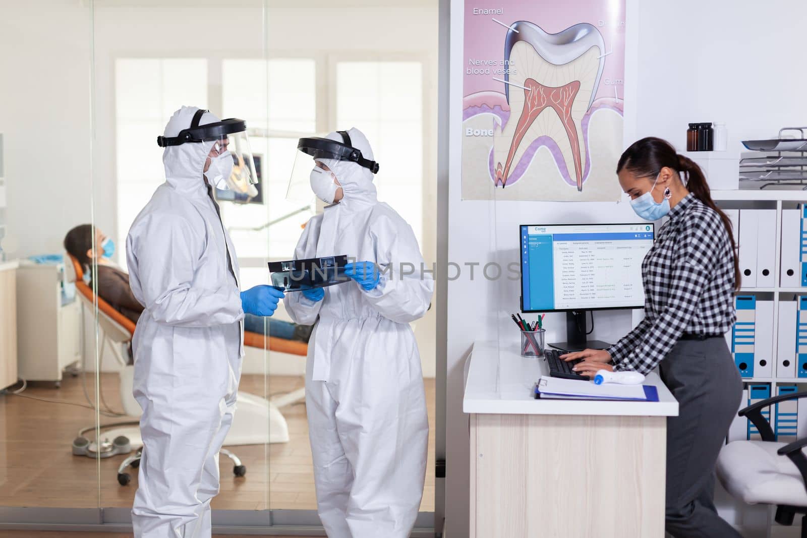 Stomatology team dressed up in ppe suit during global pandemic with coronavirus in dental reception holding patient x-ray, keeping social distancing. Receptionist with face mask as prevention.