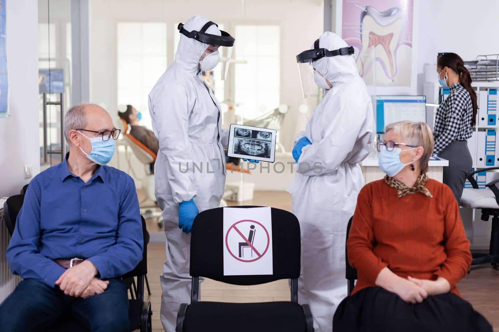 Man discussing with nurse in dental reception wearing protection suit against coronavirus, elderly patients waiting in reception keeping distance. Concept of new normal dentist visit in outbreak.