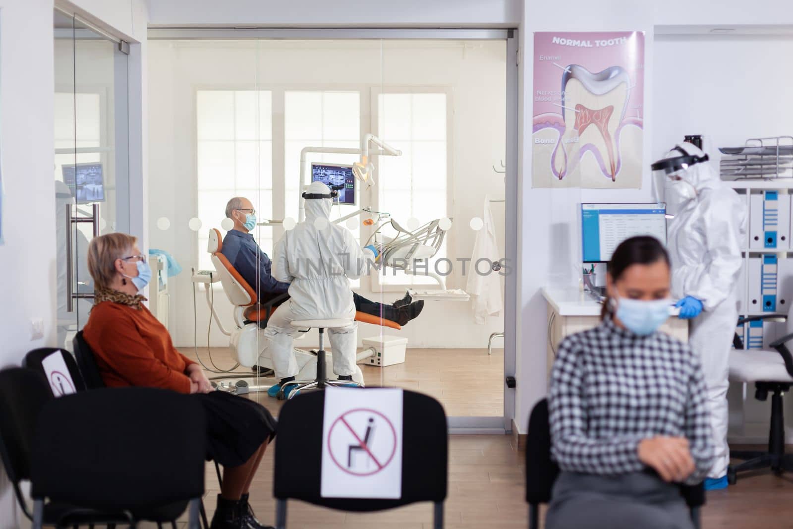 Doctor in pee suit consulting senior patient in stomatology office keeping visor, during global pandemic with coronavirus, new normal. Receptionist with face mask in dentistiry waiting area.