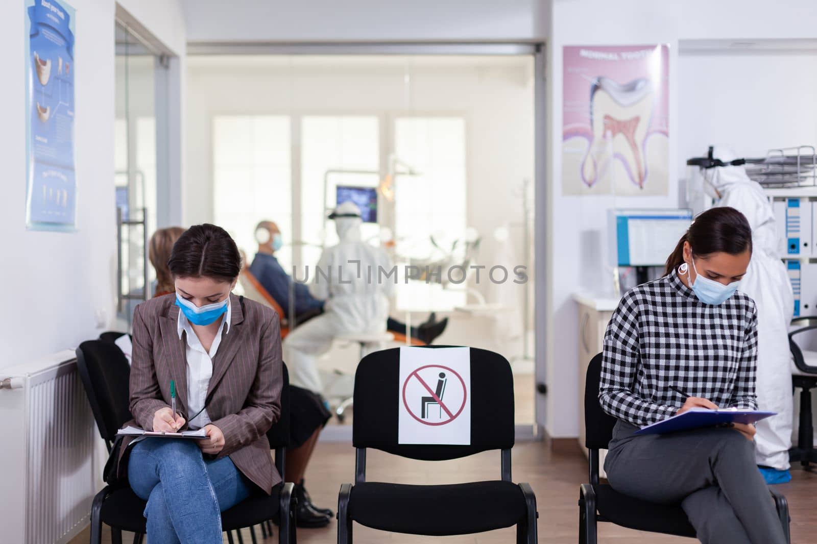 Patient with face protection mask writing on registration form in stomatomoly clinic, sitting in reception respecting social distance. Dentistiry doctor receptionist dressed in ppe suit.