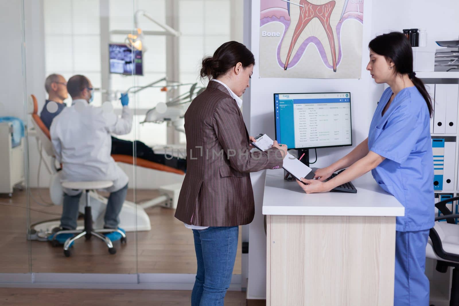 Young lady asking informations filling in stomatological form while patients talking sitting on chair in waiting area. People speaking in crowded professional orthodontist reception office.