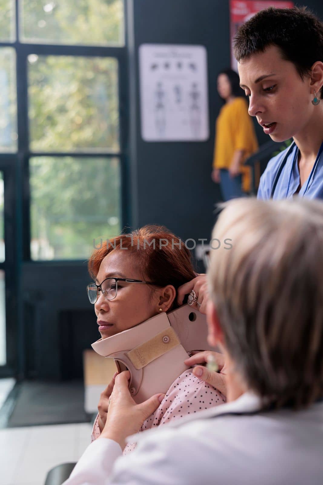 Medical staff helping injured woman to remove cervical neck collar during examination in hospital waiting area. Nurse and traumatologist medic giving support to old patient, rehabilitation therapy.