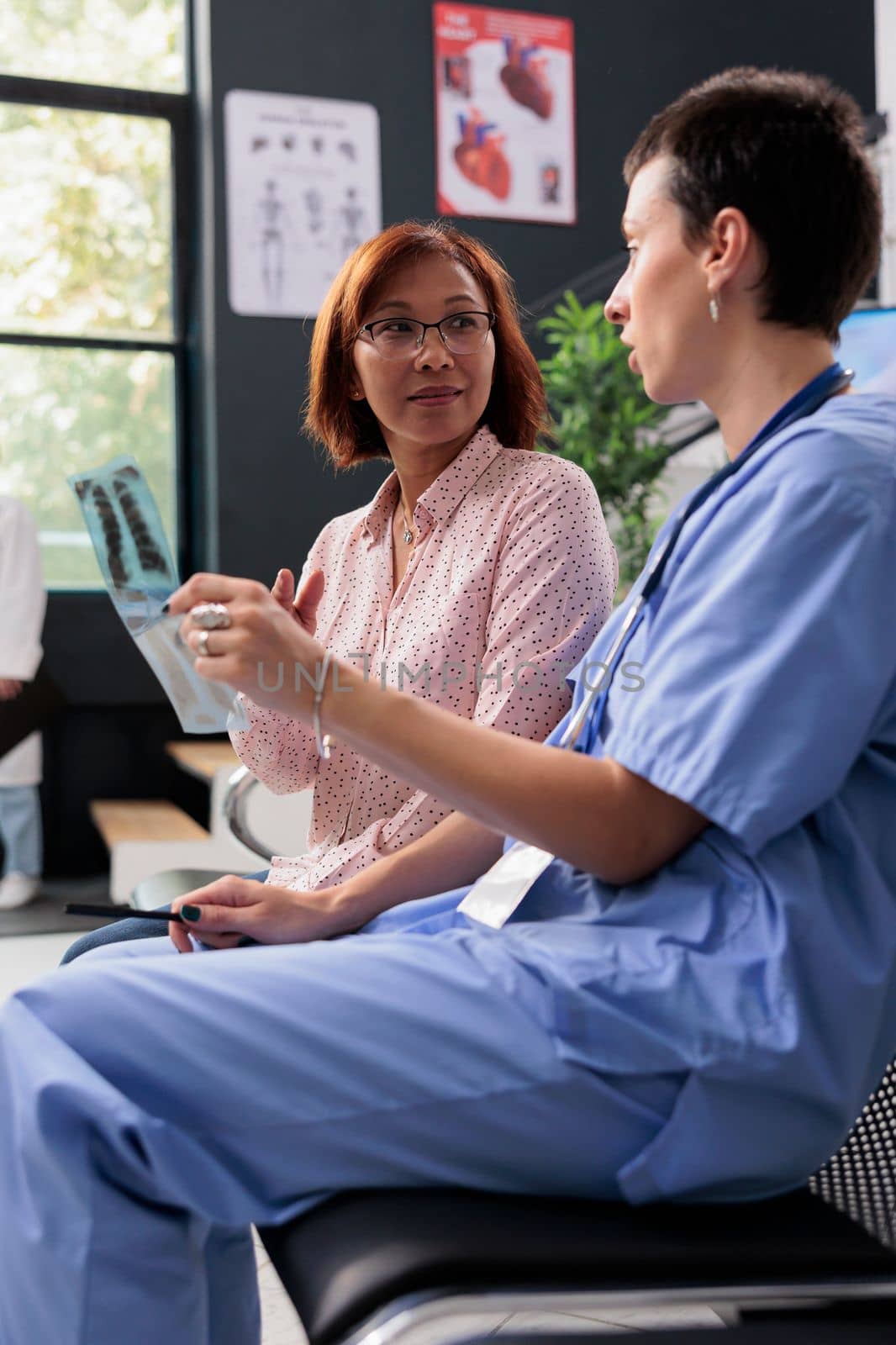 Medical assistant holding lungs xray scan explaining expertise to senior patient in hospital waiting area. Nurse discussing healthcare treatment during clinical consultation