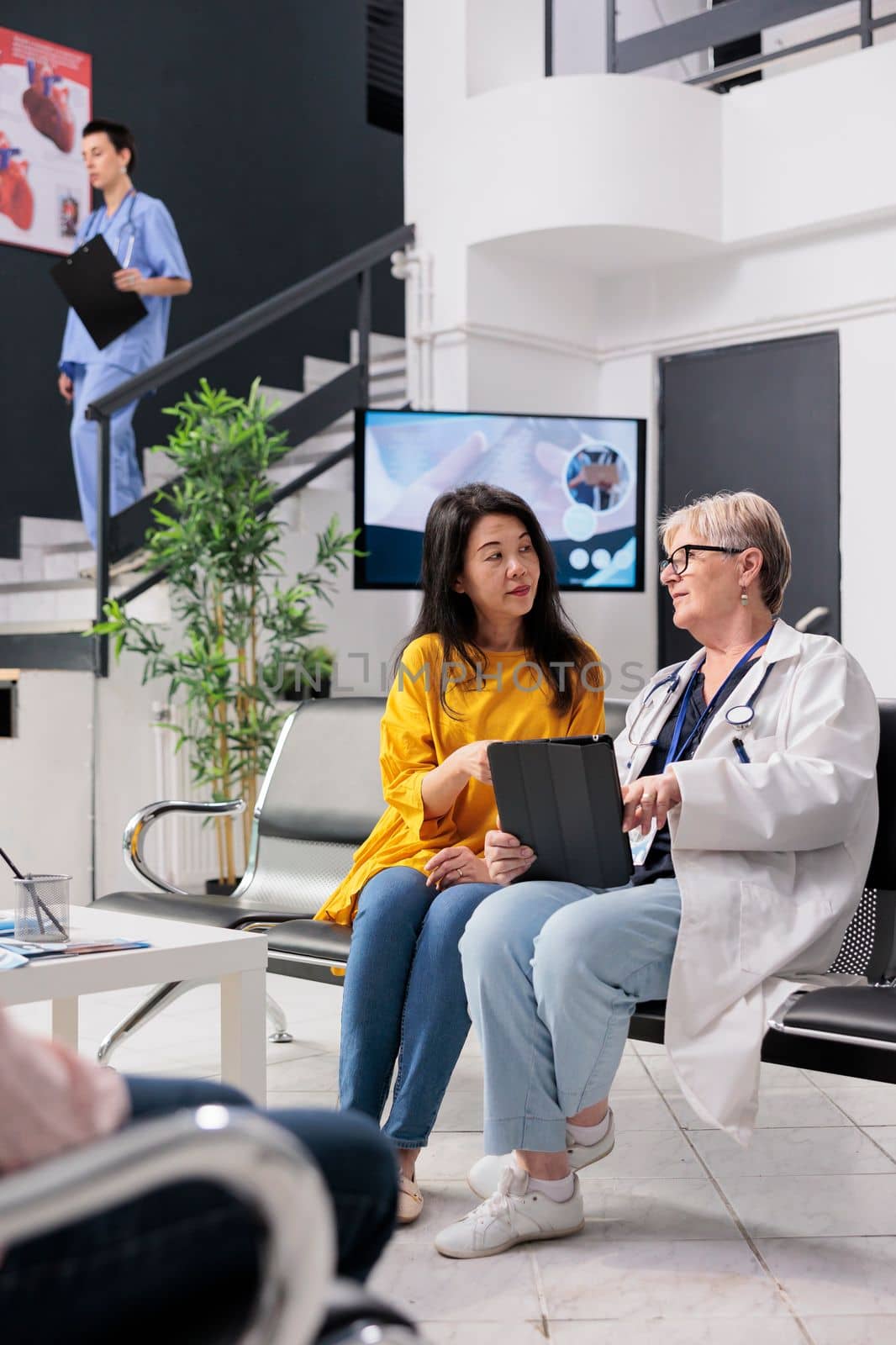 Old physician consulting asian patient at health center, analyzing disease report using digital tablet. Senior doctor talking to person with disease diagnosis at consultation in hospital waiting room