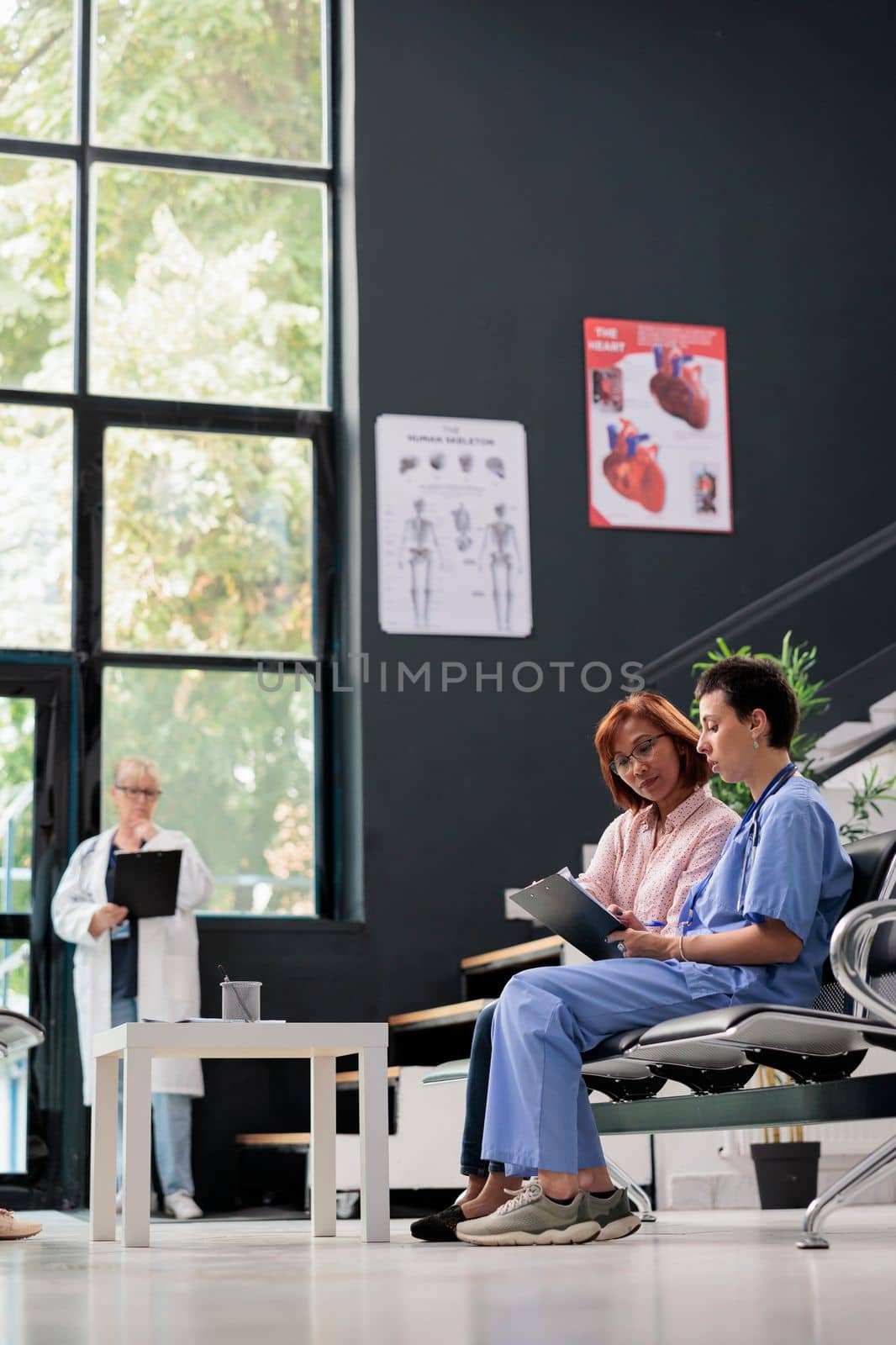 Medical asisstant consulting senior patient in hospital waiting area, filling report papers explaining healthcare treatment. Nurse doing checkup examination with asian woman in lobby.