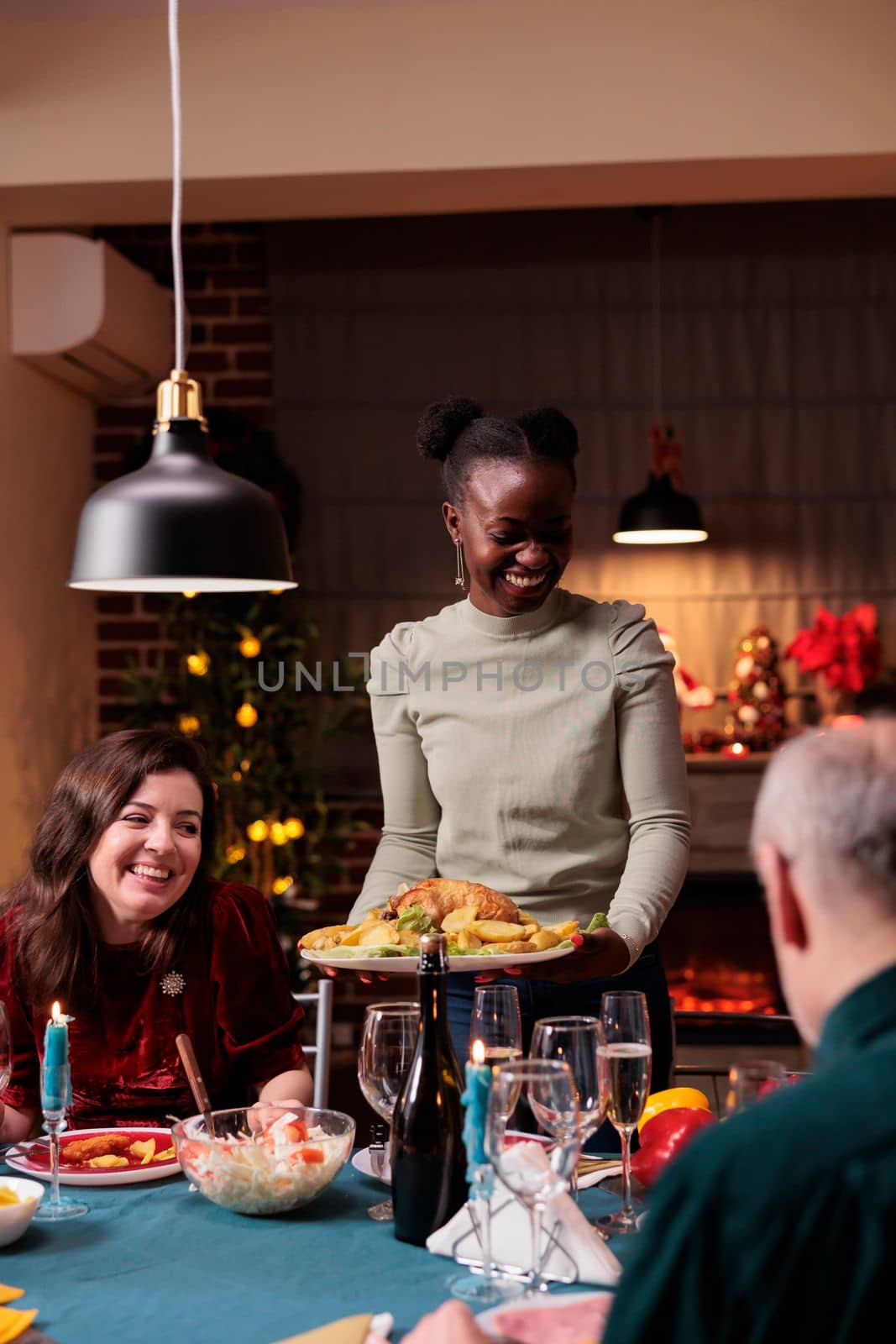 Happy family celebrating christmas, smiling woman holding traditional festive dish. Diverse people xmas celebration, winter holiday home evening party, served dinner table