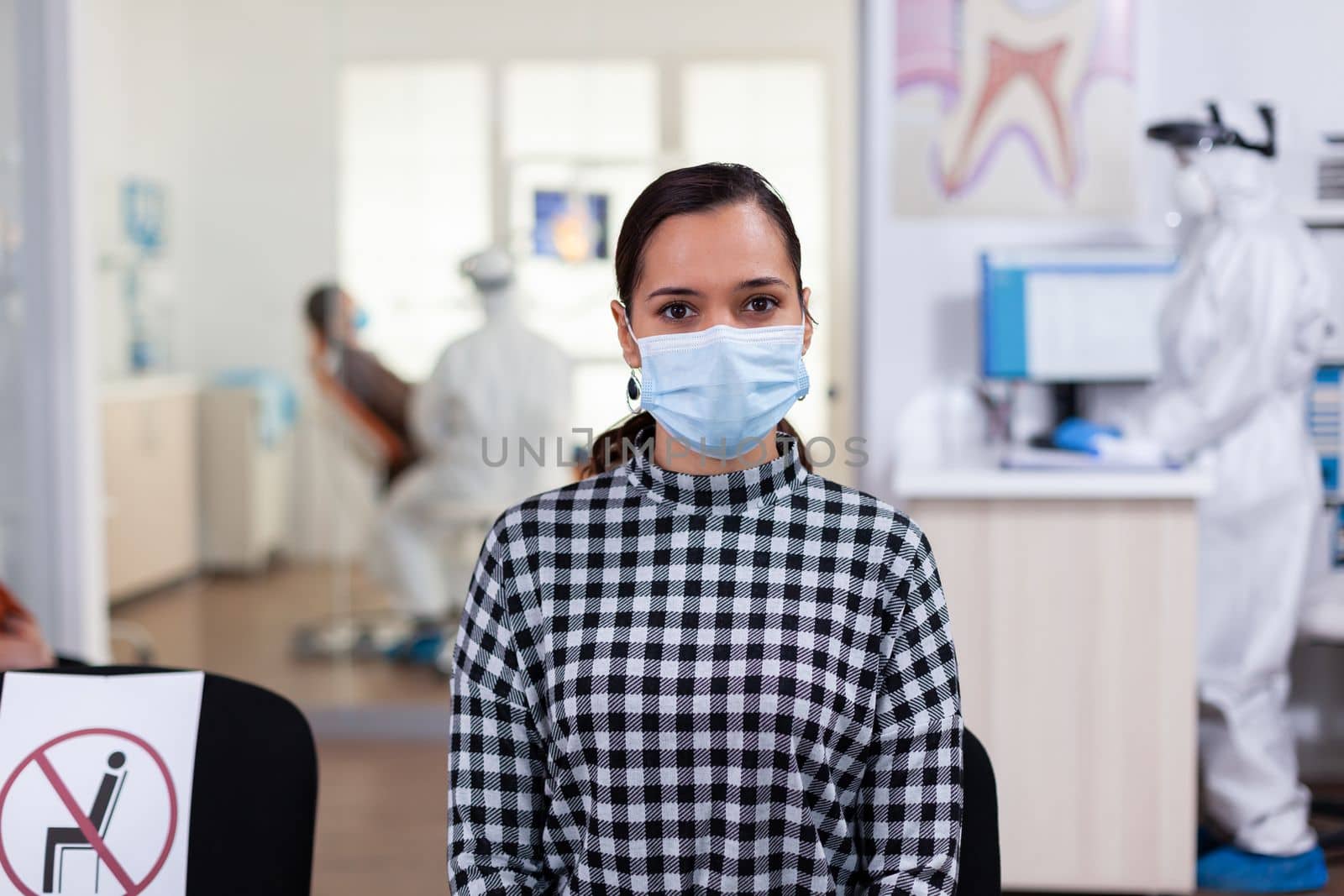 Portrait of woman in dental office looking on camera wearing face mask sitting on chair in waiting room clinic while doctor working. Concept of new normal dentist visit in coronavirus outbreak.