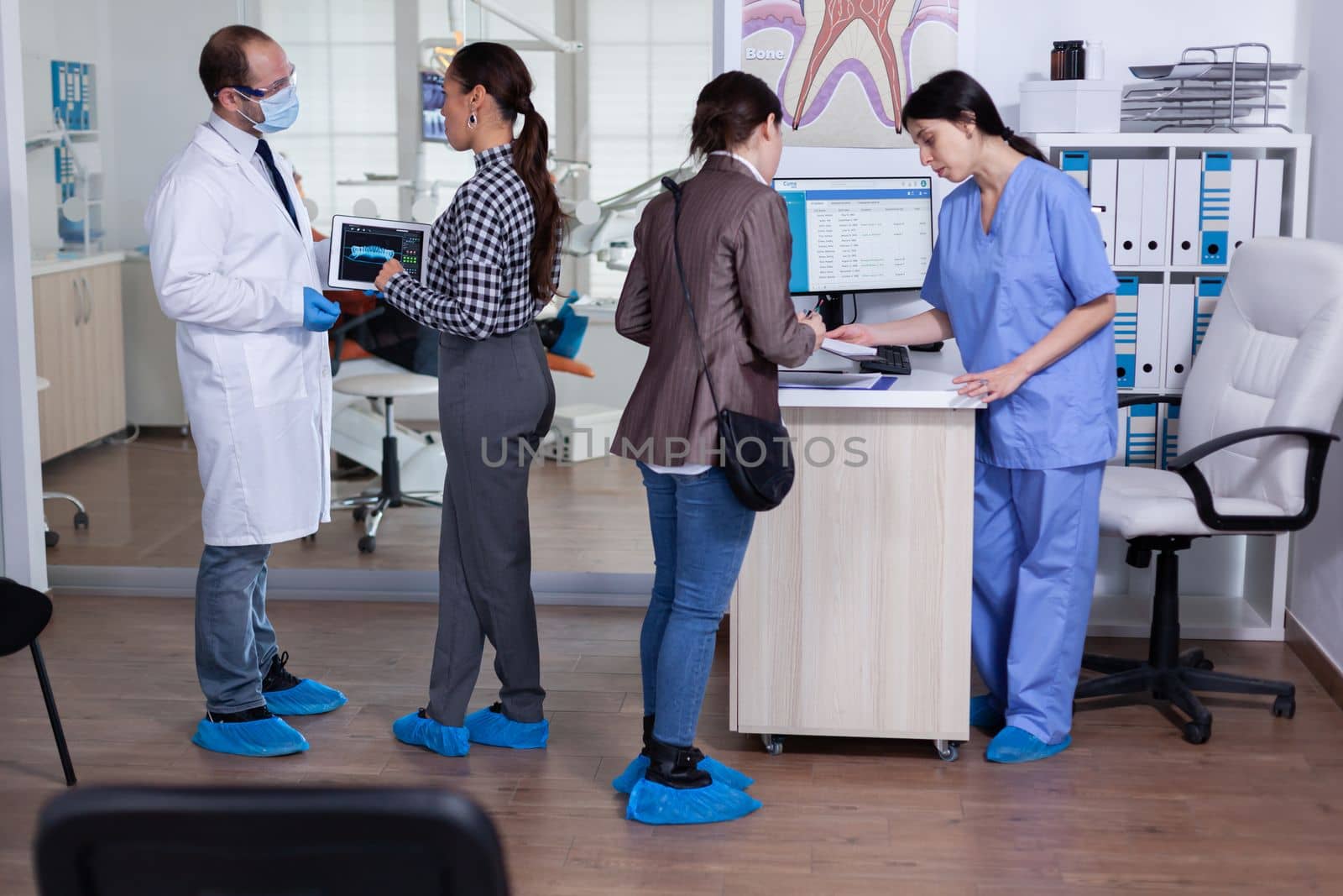 Dentist with face mask pointing at digital teeth radiography x-ray to patient in clinic waiting area. Receptionist writing on computer people appointments.