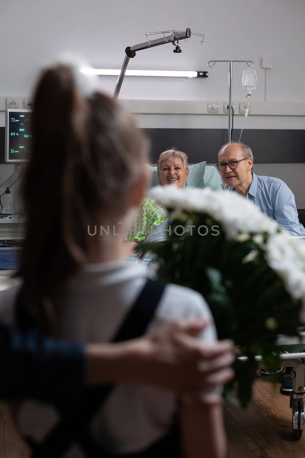 Smiling grandmother watching granddaughter arriving for visit at hospital room, holding flowers bouquet. Elderly couple happily greeting relatives in geriatric clinic room.