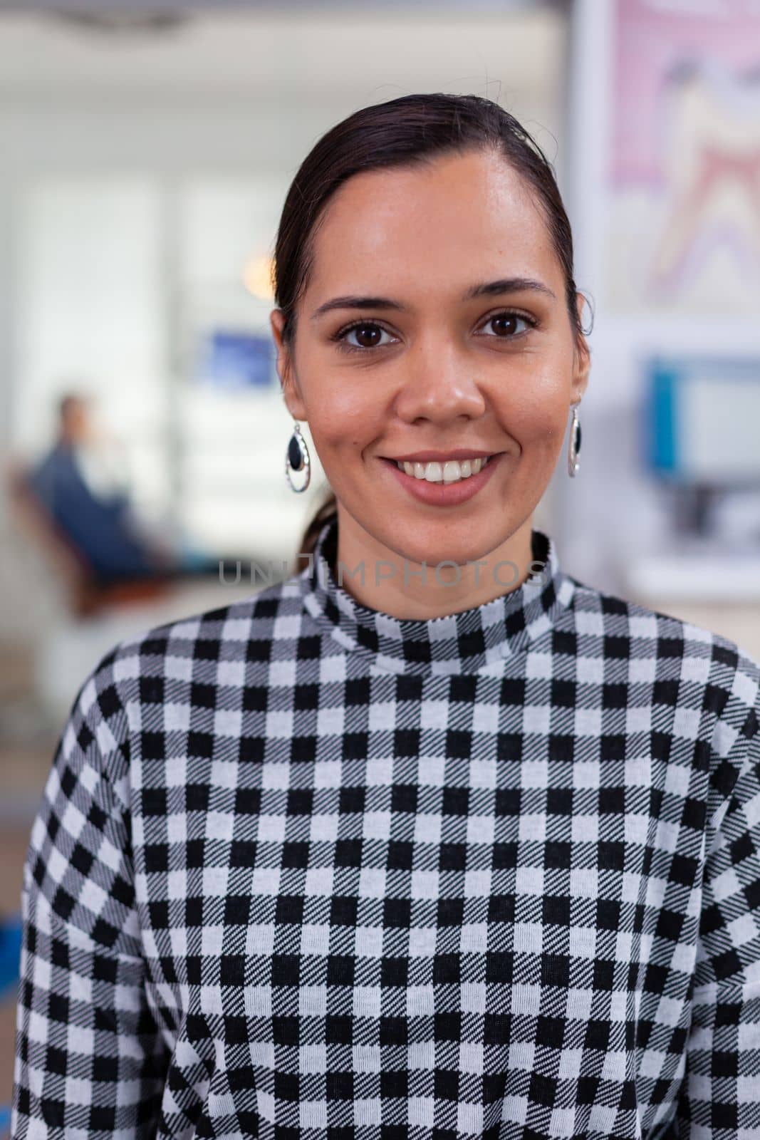 Portrait of smiling woman patient looking on camera sitting on chair in waiting room of stomatological clinic. Stomatologist assistant typing on pc in dental office while doctor working in background.