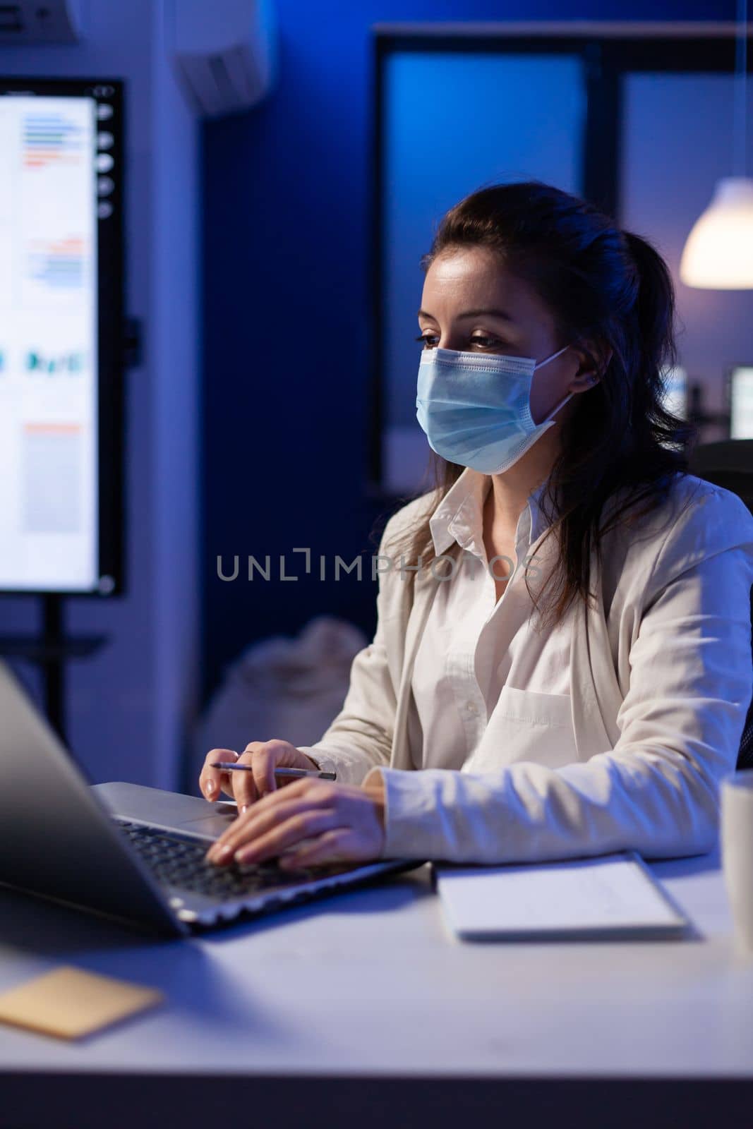 Manager woman with face mask working overtime in new normal business office looking at team project on professional laptop. Analysing financial documents sitting at desk during global pandemic