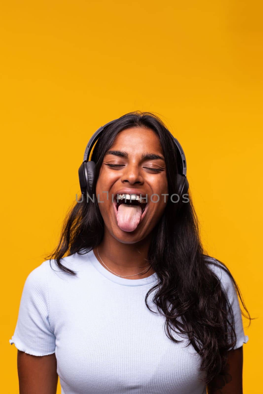 Vertical portrait of happy young Indian woman wearing wireless headphones showing tongue to camera. Yellow background. by Hoverstock
