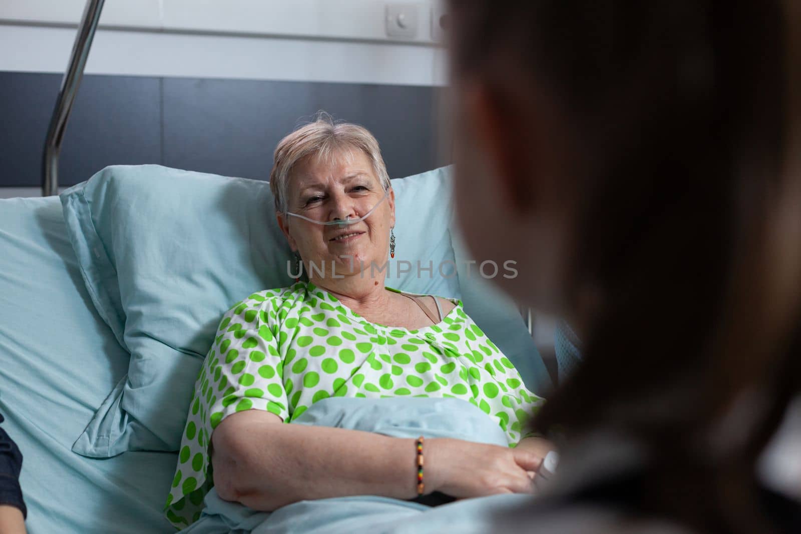 Elderly woman talking to unrecognizable girl. Close up of happy grandmother being visited by granddaughter at geriatric hospital room. Smiling old lady talking to female relative in sanatorium bed.