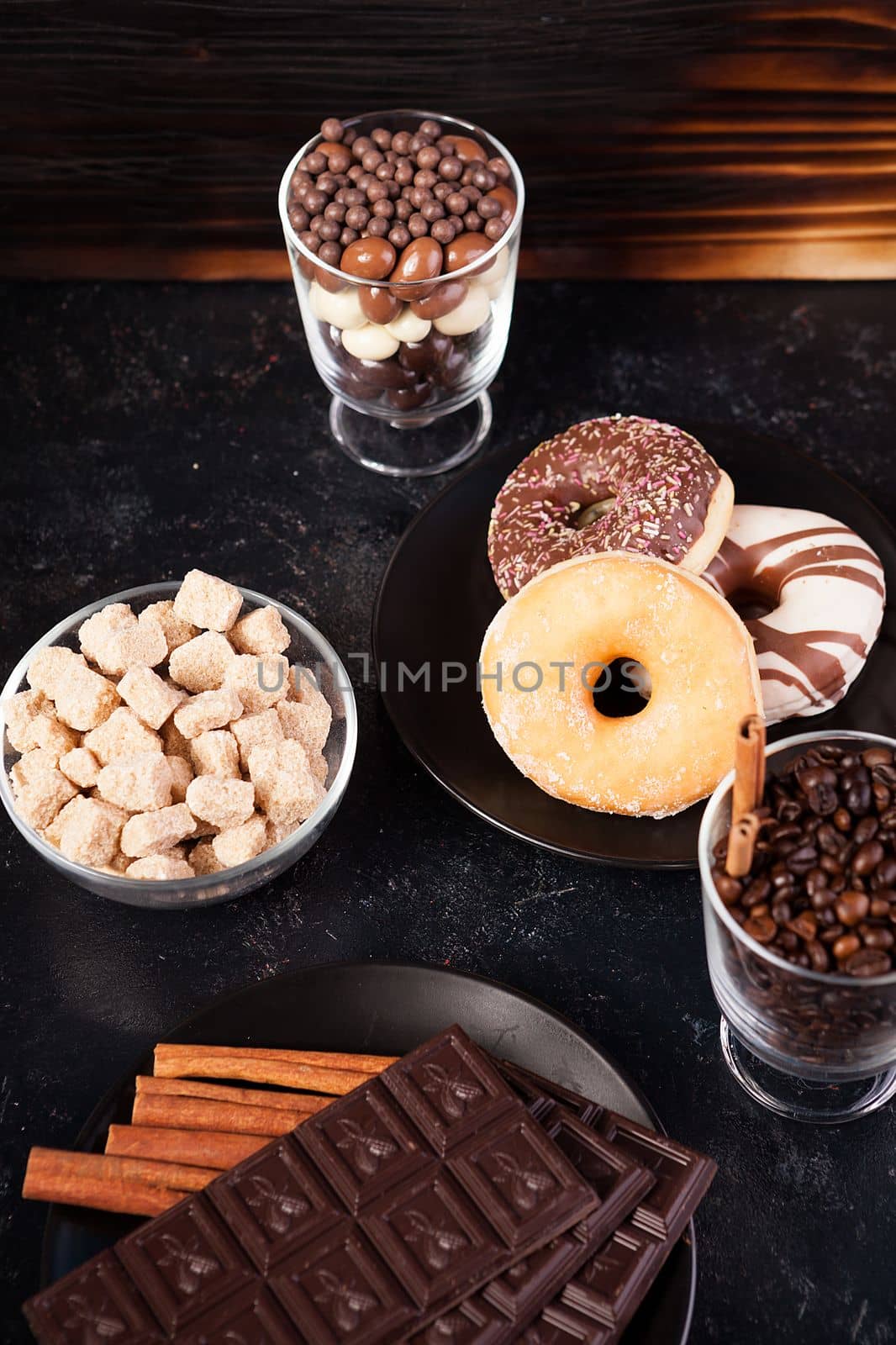 Glass with coffee beans next to chocolate tablets, donuts, brown sugar and other glass with peanuts in chocolate on dark wooden background