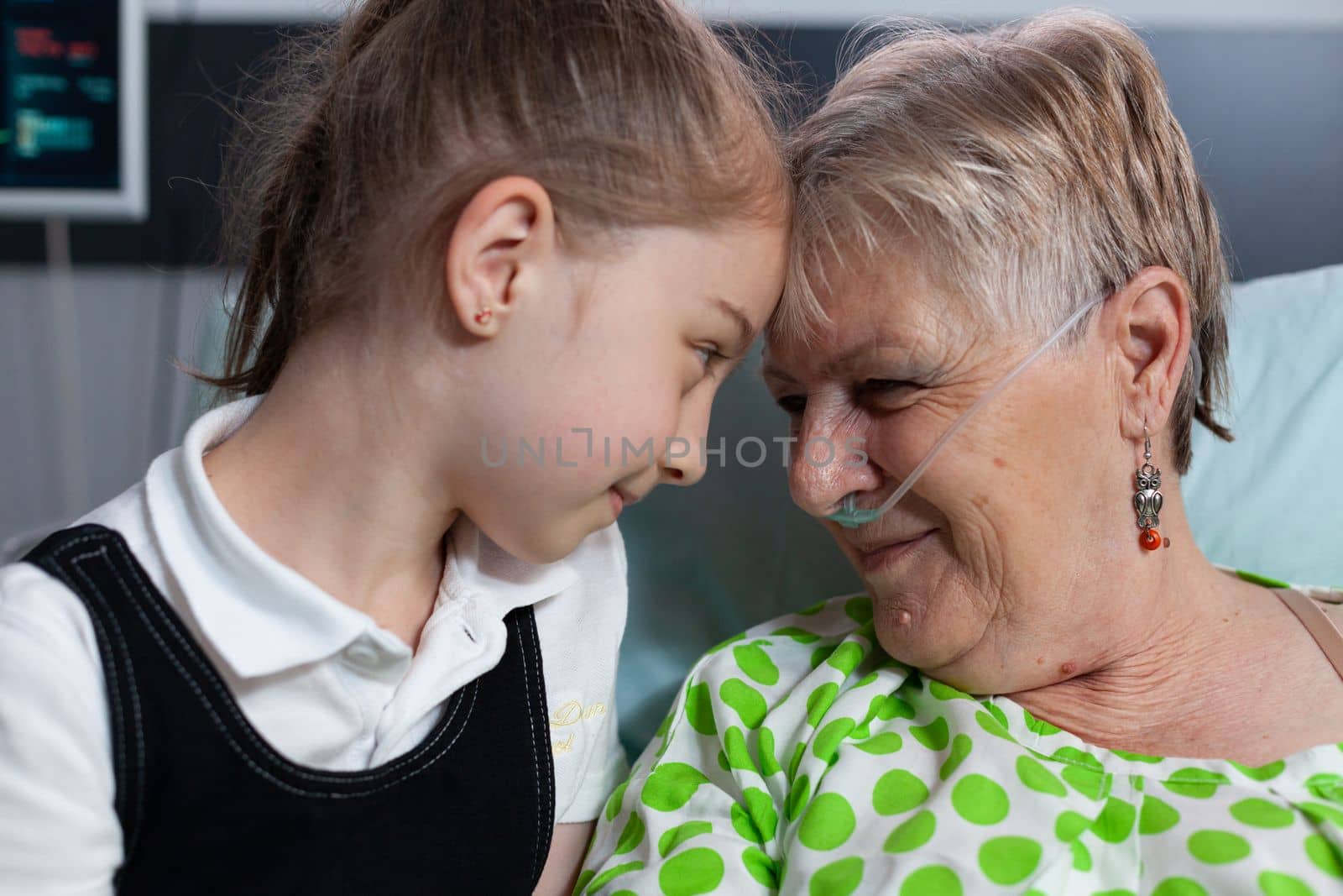 Elderly woman lying in hospital bed with breathing aids showing affection to little girl. Grandmother and granddaughter bumping heads in love at medical clinic for elderly people recovery room.