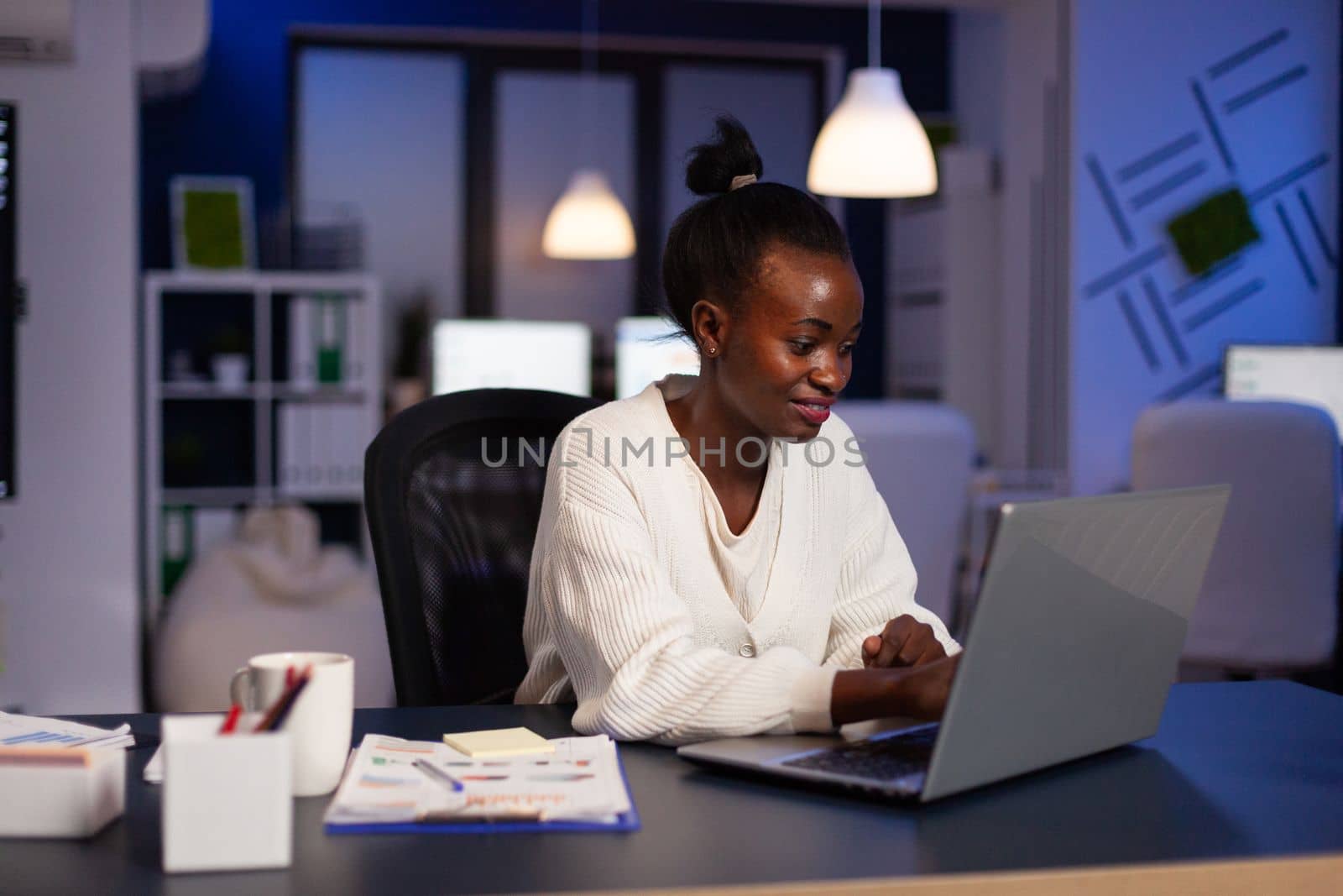 Smiling african american businesswoman planning management annual graph typing company strategy on laptop computer late at night in startup office. Black manager browsing corporate information