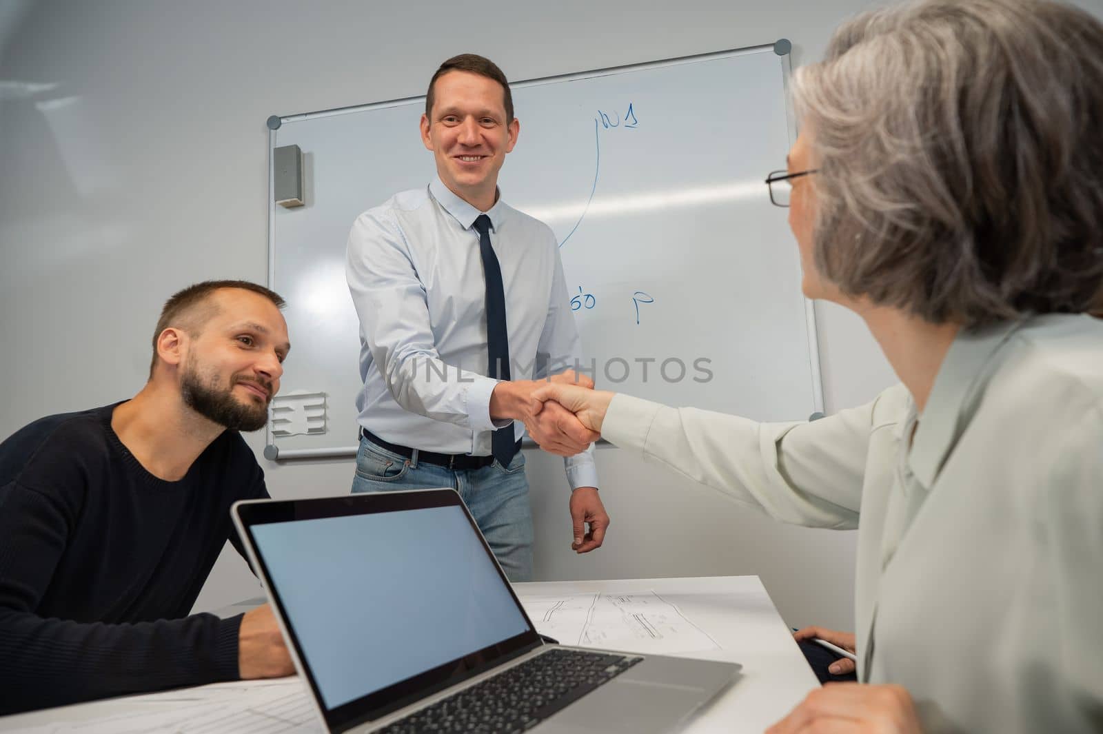 The boss makes a presentation to subordinates at the white board. Caucasian man shaking hands with middle aged woman