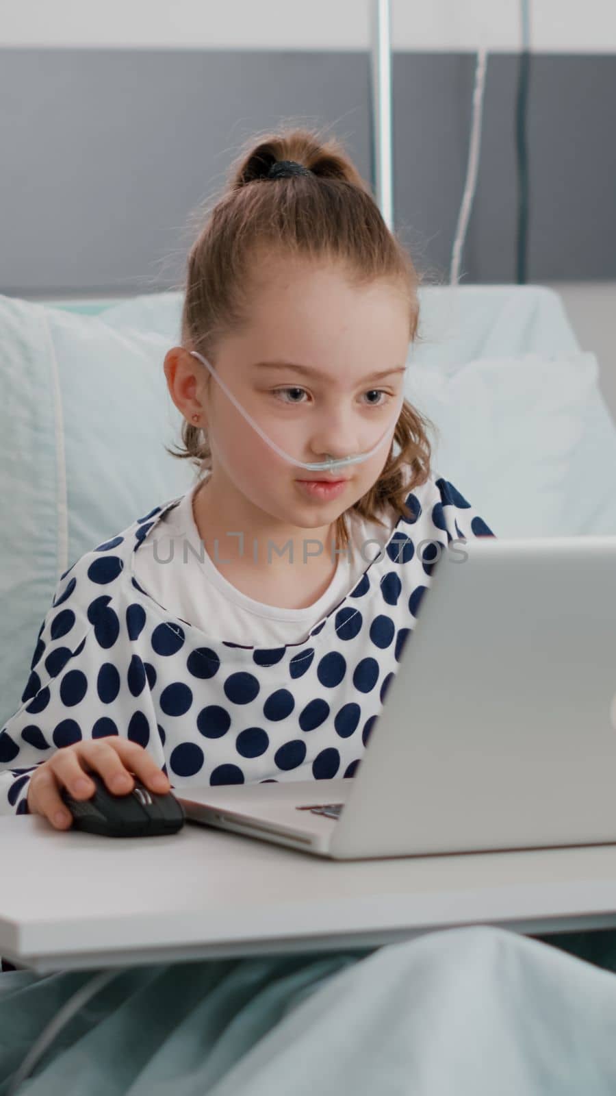 Hospitalized sick girl playing online videogames using laptop computer during recovery examination in hospital ward. Child sitting in bedwearing oxygen nasal tube having breathing sickness