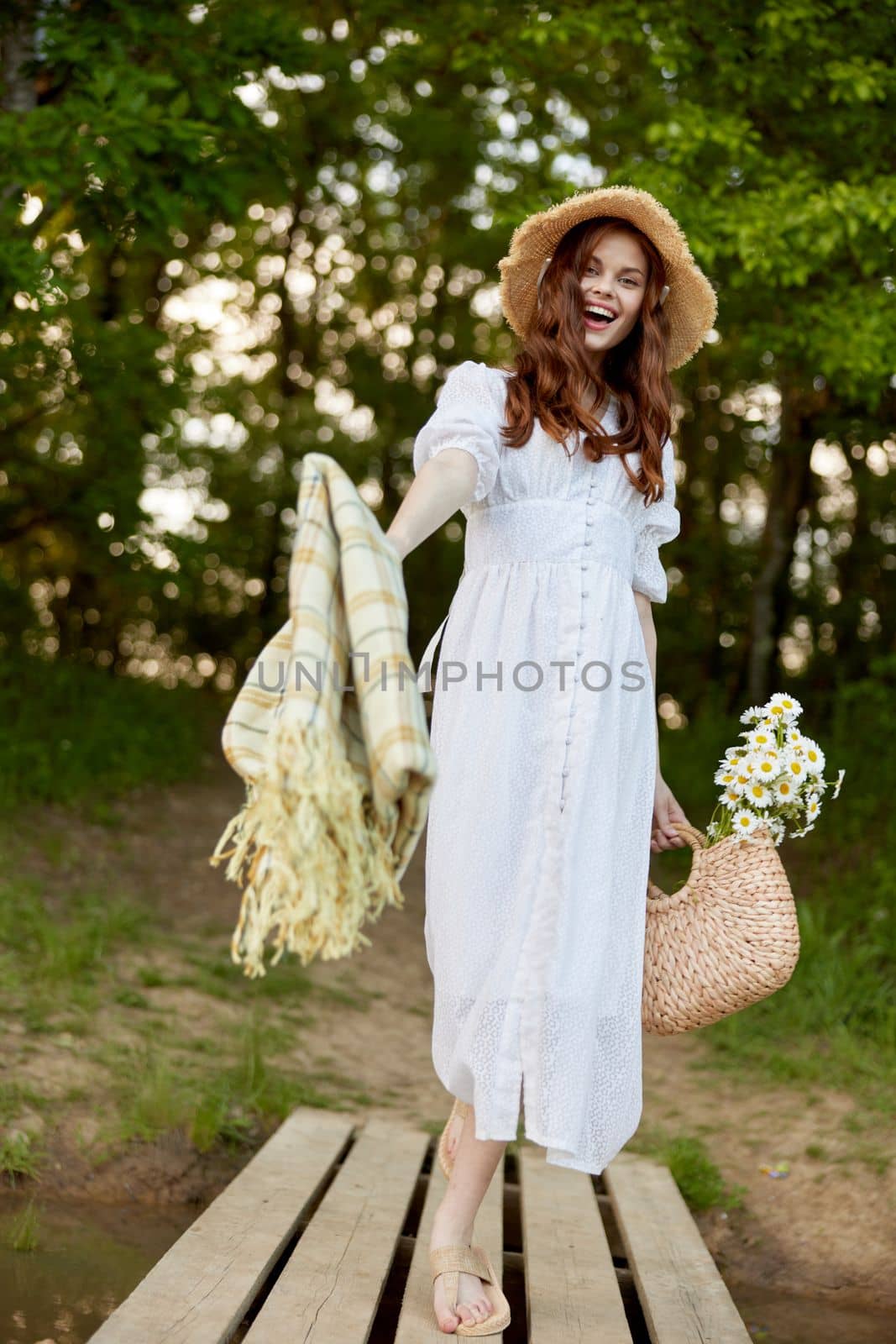 a cute woman in a long summer dress with a plaid in her hands and a wicker hat goes to rest on the pier. High quality photo