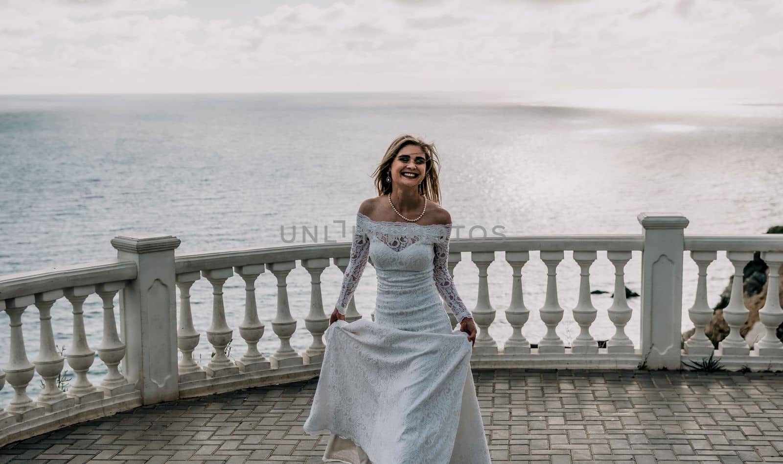 Romantic bride, blond girl in white wedding dress with open shoulders posing on open terrace with backdrop of the sea and rocks. Stylish young woman standing on terrace and looking on ocean at sunset by panophotograph