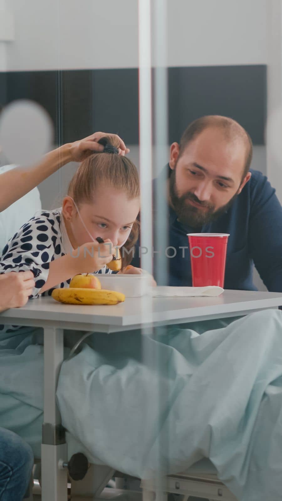 Family sitting with sick little daughter while eating healthy nutrition food during recovery examination in hospital ward. Child patient wearing oxygen nasal tube resting in bed having meal lunch