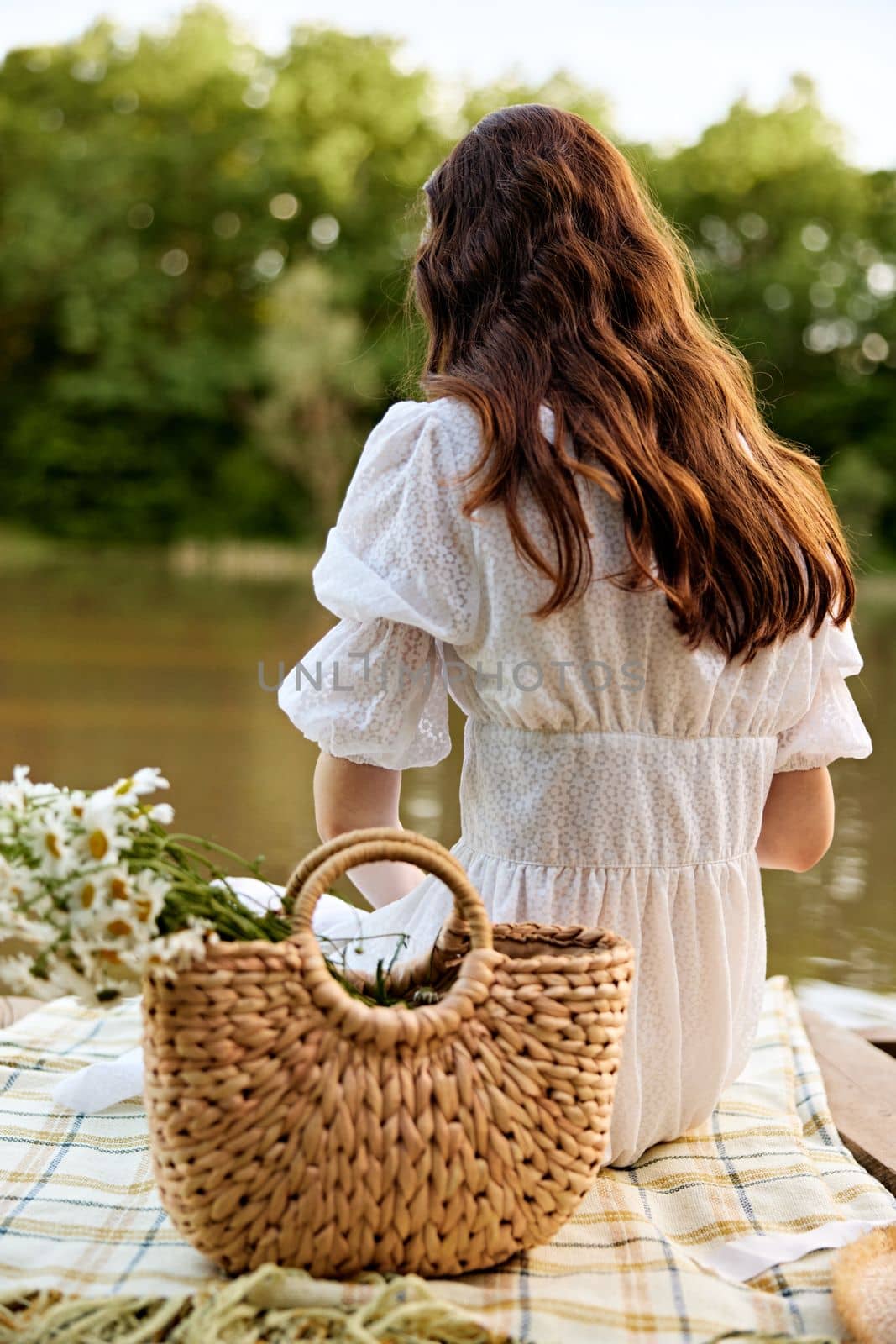 close-up photo of a woman sitting with her back to the camera on the shore of a lake with a wicker bag full of daisies by Vichizh