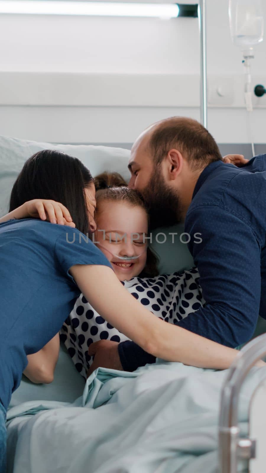 Happy parents visiting sick daughter hugging child during disease examination in hospital ward. Hospitalized kid wearing oxygen nasal tube resting in bed after suffering recovery surgery