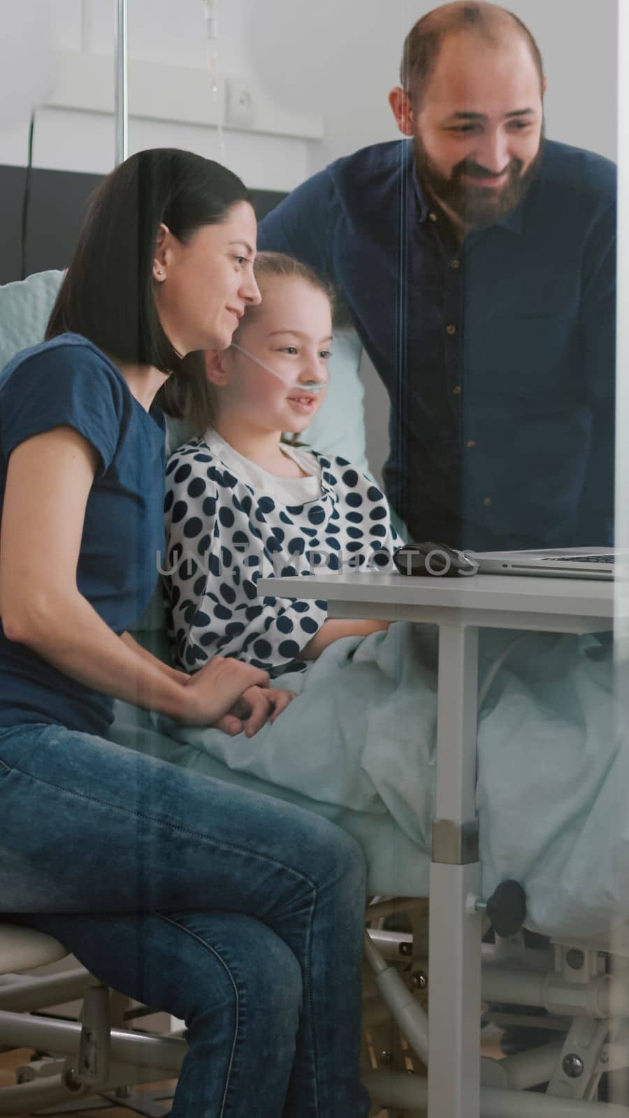 Family greeting remote friends during online videocall telecommunication using laptop computer in hospital ward. Sick daughter resting in bed recovering after medical surgery waiting disease treatment