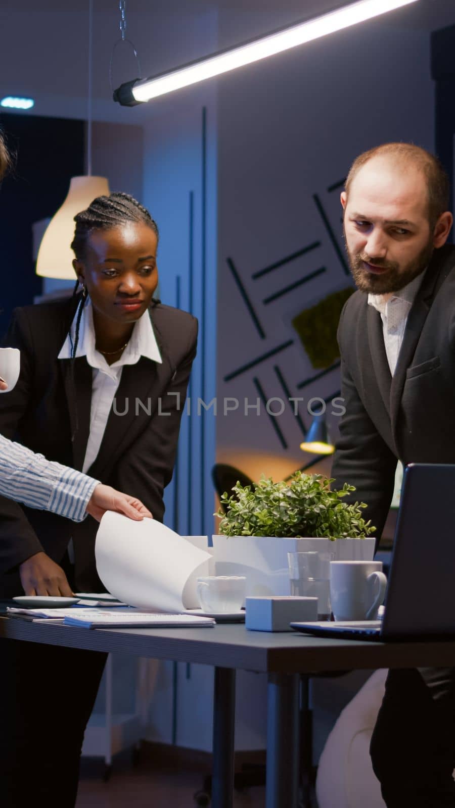 Business diverse teamwork solving management strategy analyzing company graphs using computer while standing at conference table. Multi ethnic businesspeople overworking in meeting room in evening