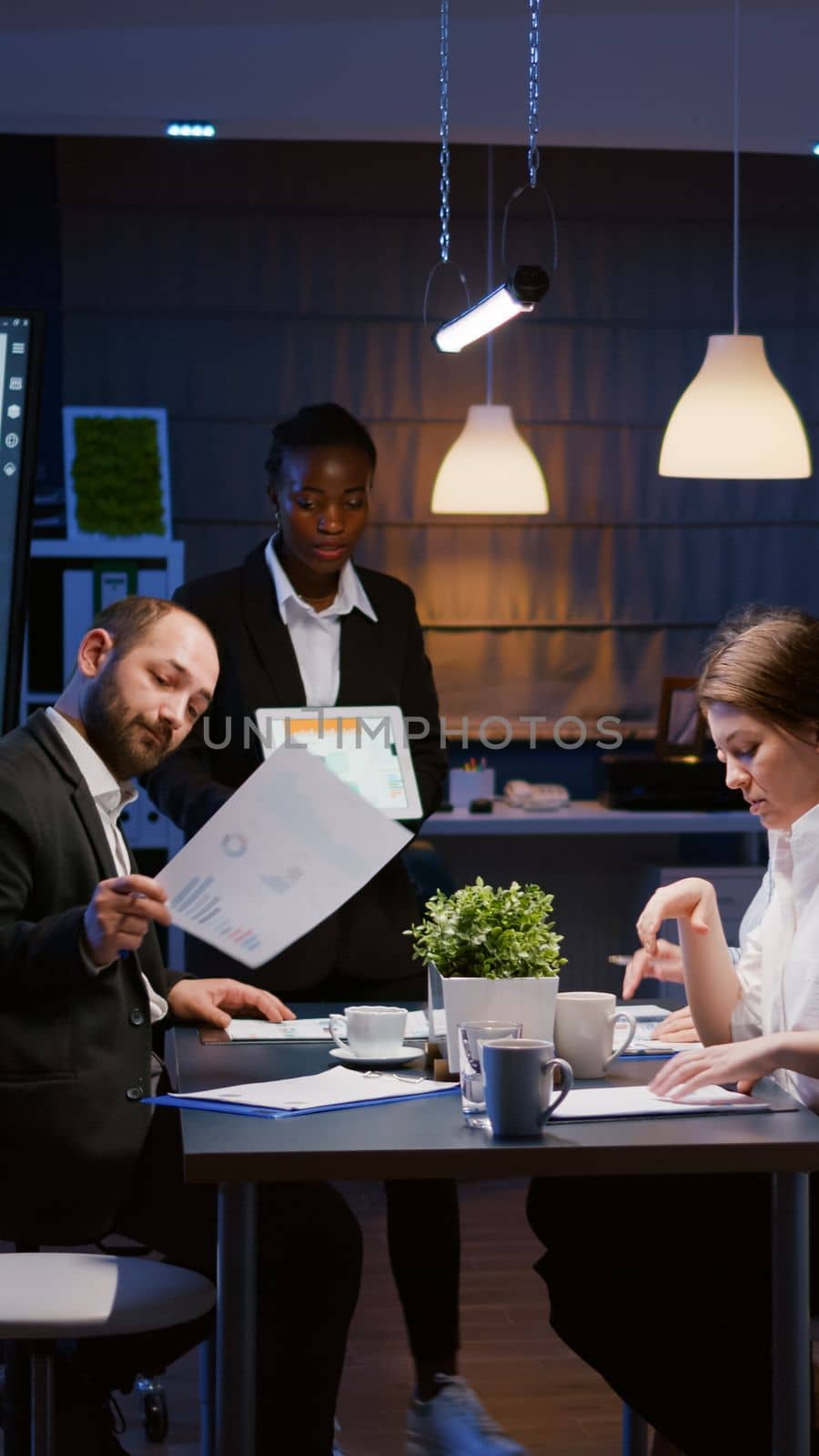 Focused workaholic entrepreneur woman with dark skin explaining management strategy using tablet. Business diverse multi-ethnic teamwork overworking in company office meeting room late at night