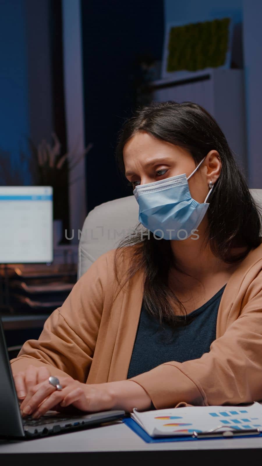 Workaholic businesswoman with face mask against covid19 working in startup office analyzing economic strategy late at night. Exhausted manager remains alone in company room after collegue left