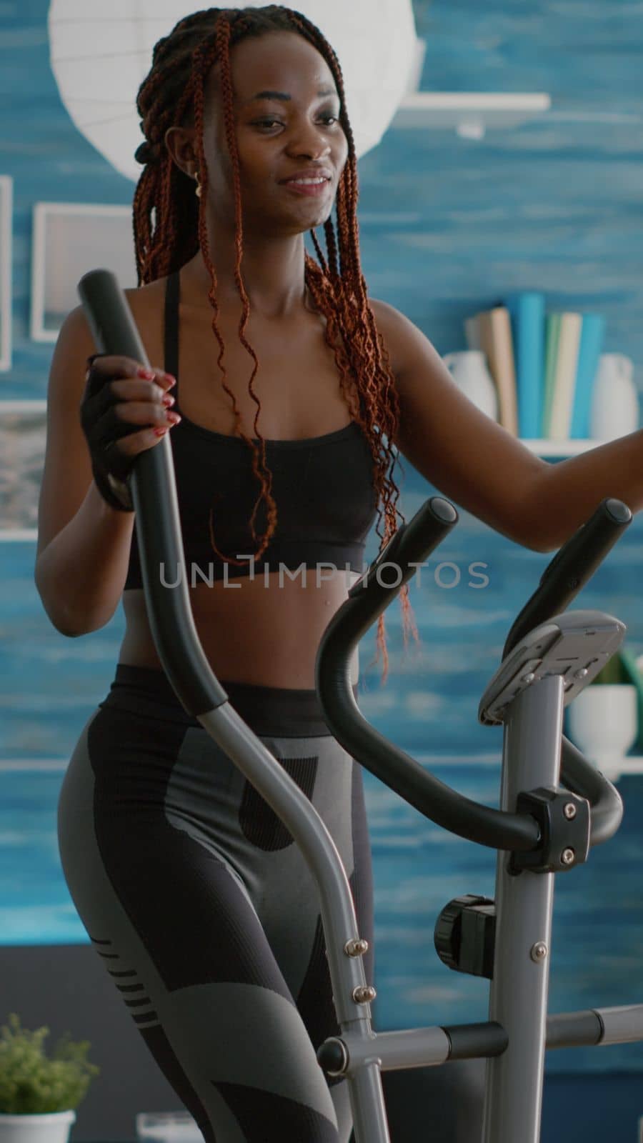 Black athletic woman doing cardio training on elliptical bike in living room by DCStudio