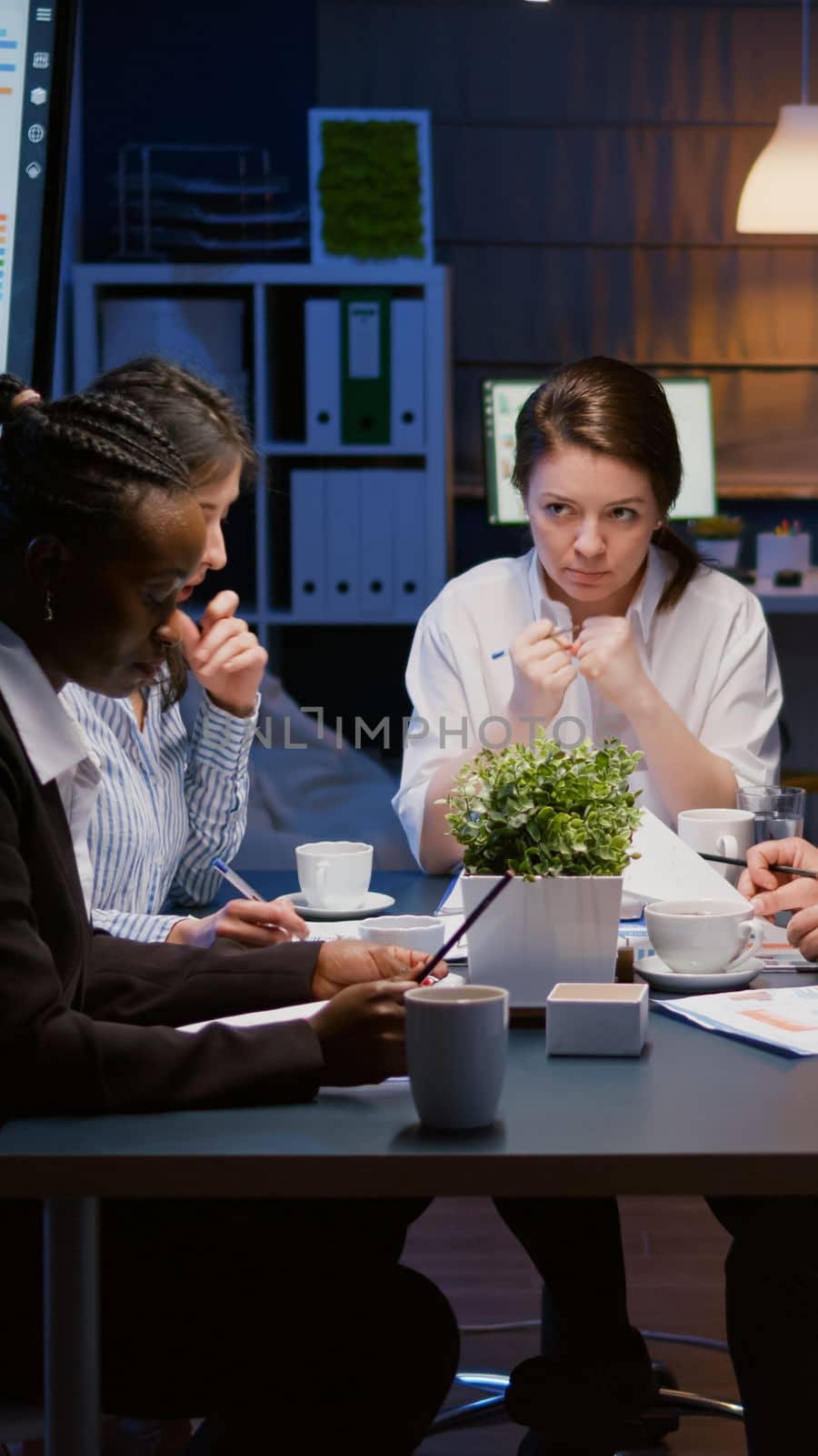 Diverse multiethnic business teamwork overworking in office meeting room analyzing financial graphs paperwork late at night. Coworkers brainstorming project ideas solving company problem