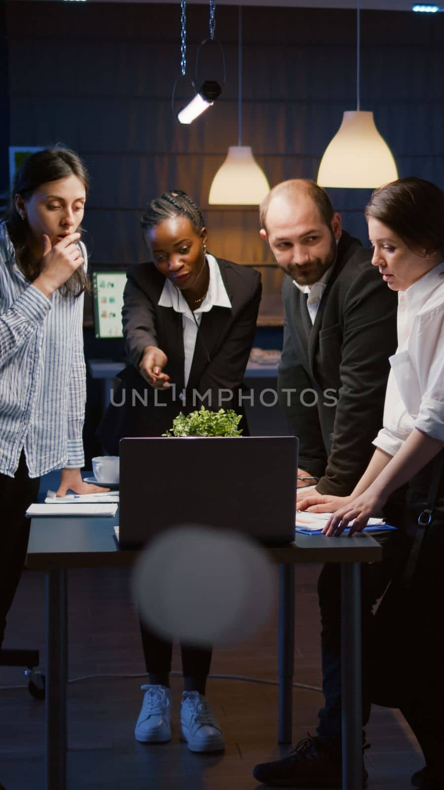 Multi ethnic businesspeople analyzing management presentation using laptop computer overworking in office meeting room late at night. Diverse teamwork brainstorming overtime business solution