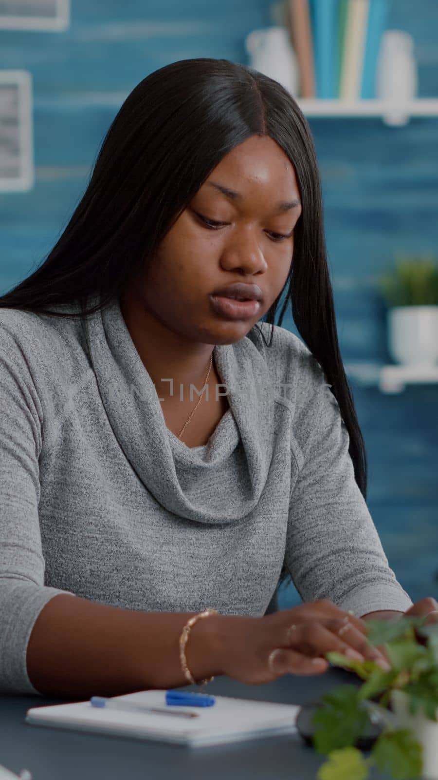 Black student sitting at desk writing school homework on notebook during online courses education. Young woman browsing communication project on computer in living room typing webinar notes